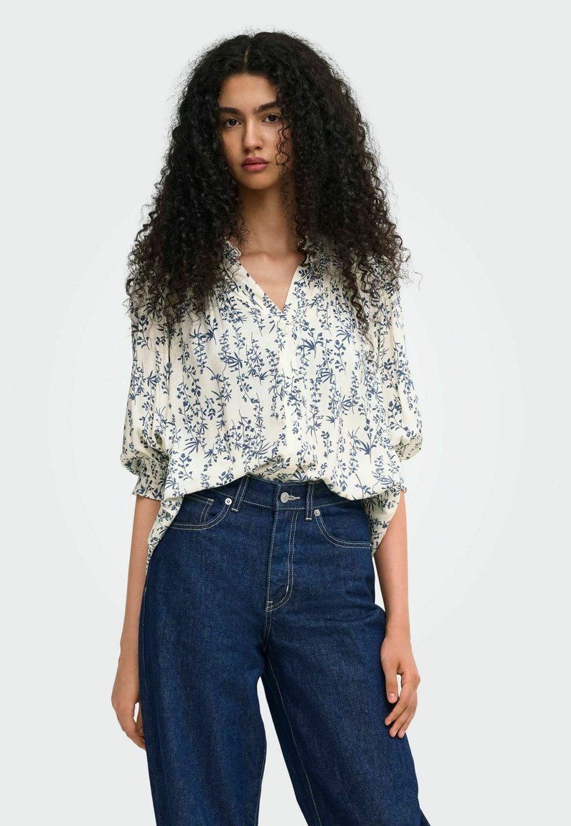 Femme aux longs cheveux bouclés portant un chemisier blanc à imprimé floral bleu et un jean taille haute bleu foncé sur fond blanc.