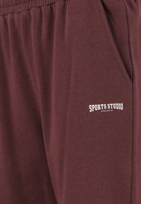 Bordauxfarbene Jogginghosen aus weicher Baumwolle, ausgestattet mit einem elastischen Bund und Seitentaschen, mit einem weißen Logo, das "SPORTS STUDIO ATHLETICA" liest.