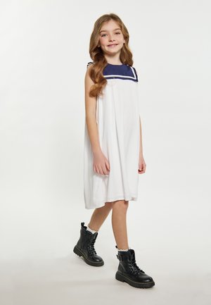 Weißes ärmelloses Kleid mit einem marineblauen Kragen und Besatz, kombiniert mit schwarzen schnürbaren Kampfstiefeln. Das Kleid hat ein fließendes Design und reicht bis zur Mitte der Waden.