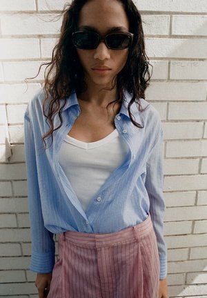 Femme aux longs cheveux bouclés portant des lunettes de soleil noires, une chemise rayée bleu clair sur un haut blanc et un pantalon rayé rose taille haute devant un mur en briques blanches.