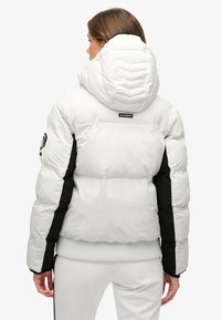 Chaqueta blanca acolchada con paneles laterales negros, cuello alto y capucha ajustada. Presenta textura acolchada y logo en la espalda.