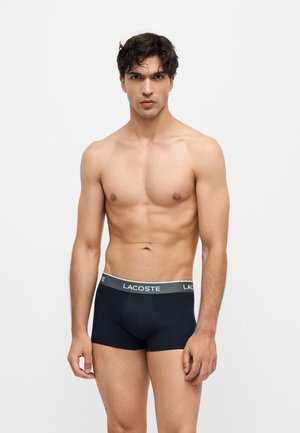 Uomo muscoloso con capelli scuri in piedi di fronte, indossando boxer Lacoste neri su uno sfondo chiaro e uniforme.