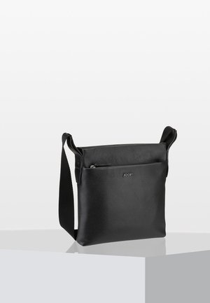 Sac bandoulière en cuir noir texturé avec poche zippée à l'avant et sangle en tissu réglable, posé sur un socle blanc.