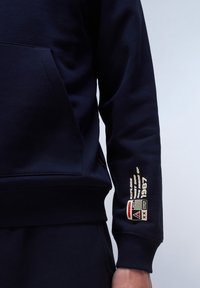 Sudadera azul marino con bolsillo frontal, puños y dobladillo acanalados. Presenta un logo gráfico y detalle de texto en la manga izquierda. Textura de tela suave.