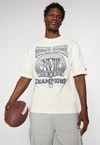 Biele tričko s krátkym rukávom v bielej farbe s grafickým dizajnom Super Bowl Raiders v sivej farbe, v kombinácii so sivými teplákmi, držiac hnedý futbal.