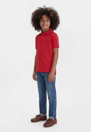 Bambino sorridente con capelli ricci che indossa una polo rossa, jeans blu risvoltati e scarpe di pelle marrone, in piedi su uno sfondo bianco uniforme.