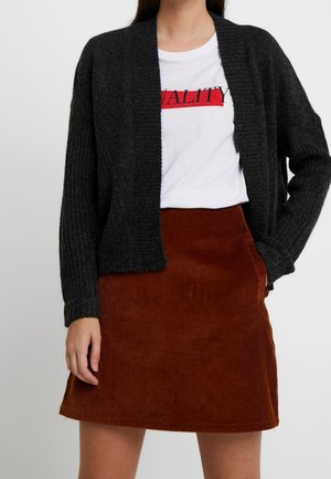 Femme portant un T-shirt blanc avec le texte "QUALITY", un cardigan ouvert gris charbon et une jupe marron en velours côtelé coupe trapèze avec poches.