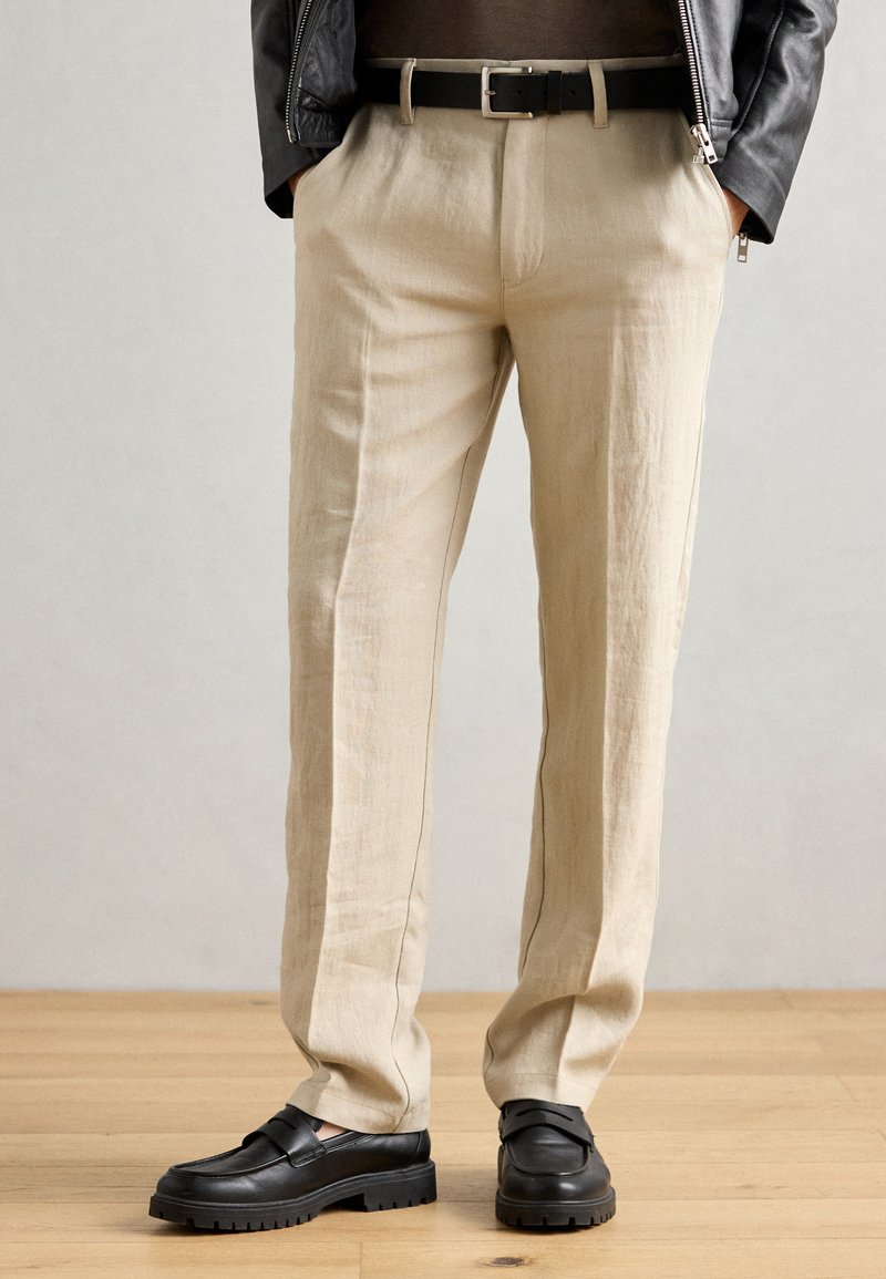 Pantalon en lin beige avec une coupe droite, présentant un pli, associé à des chaussures noires à enfiler avec des semelles épaisses. Une ceinture noire accentue la taille.