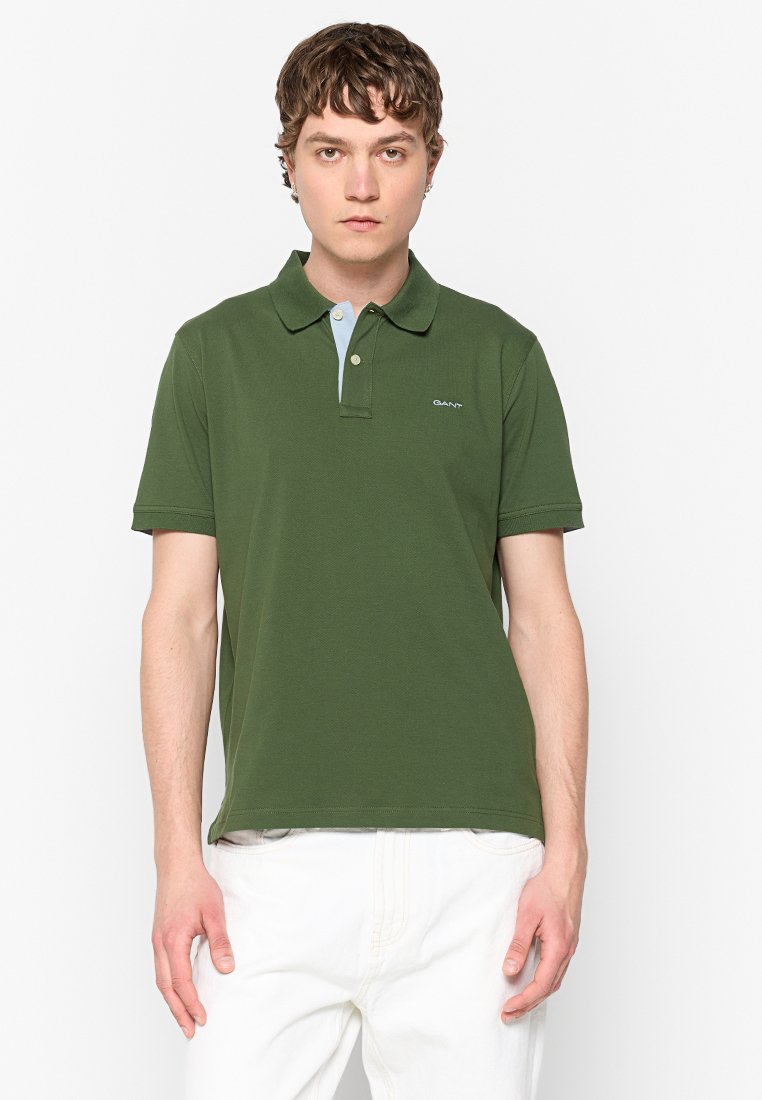Polo vert en coton, avec un col bleu clair contrastant, des manches courtes et un petit logo sur la poitrine. Pantalons blancs.