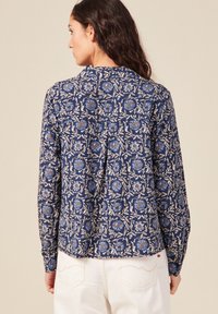 Blusa floreale navy con maniche lunghe, caratterizzata da fiori blu e arancioni su un motivo botanico crema. Dettaglio con bottoni sulla parte posteriore.