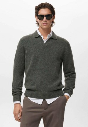 Suéter de lana verde oscuro con escote en V y cuello, puños acanalados y dobladillo. Llevado sobre una camisa blanca, combinado con pantalones grises y gafas de sol.