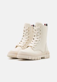 Beige enkellaarzen van gladde stof, met een vetersluiting aan de voorkant, gouden oogjes en een chunky zool met textuurdetails.