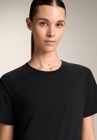 Mladá žena se rovnými tmavými vlasy staženými dozadu, oblečená v jednoduchém černém tričku s kulatým výstřihem, dívá se přímo do kamery.