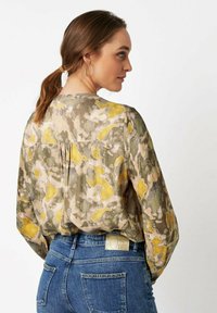 Frau mit braunem Haar im Pferdeschwanz, die eine blassgelbe, graue und grüne gemusterte Bluse sowie Blue Jeans mit Nieten an den Gesäßtaschen trägt und von hinten zu sehen ist.