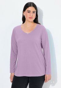 Žena s tmavými vlasy, oblečená ve stříbrných kruhových náušnicích a dlouhém světle fialovém tričku s výstřihem do V, stojící na bílém pozadí.