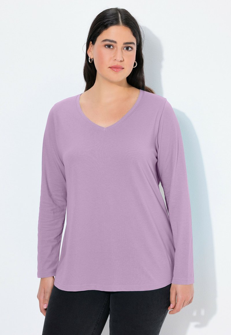 Žena s tmavými vlasy, oblečená ve stříbrných kruhových náušnicích a dlouhém světle fialovém tričku s výstřihem do V, stojící na bílém pozadí.