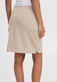 Jupe en motif chevron beige avec une légère brillance, présentant une silhouette ajustée, des poches latérales et une fente au dos pour un mouvement facilité.