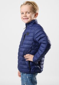 Giacca puffy blu navy con tasche con zip e colletto alto, caratterizzata da un design trapuntato e una texture liscia. Indossata sopra jeans consumati.
