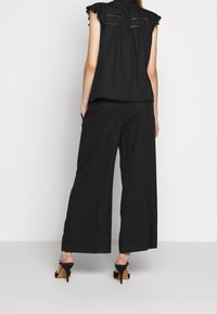 Mujer de pie con las manos en los bolsillos, vestida con una blusa negra sin mangas, pantalones anchos negros y sandalias de tacón alto negras, vista desde atrás.