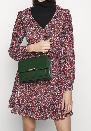 Vrouw in een lange mouw rode paisley jurk en een zwarte coltrui, die een donkergroene leren handtas met gouden kettingriem over de borst draagt.