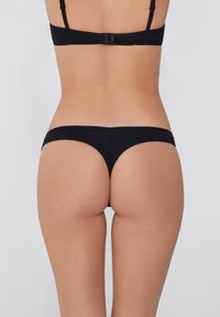 Schwarzer Tanga mit minimaler Bedeckung, glatter Textur und elegantem Design. Verstellbare Träger des passenden BHs sind hinten sichtbar.