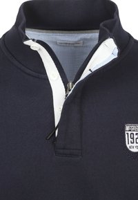 Marineblauwe polo met rits, een witte contrasterende kraag en gestreepte binnenbies. Voorzien van geborduurd logo en een zachte textuur.