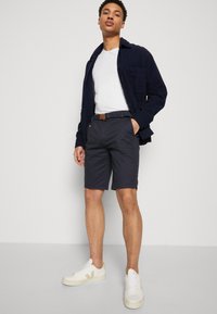 Mann står med den ene hånden i lommen iført marineblå shorts, hvit t-skjorte, marineblå jakke, brun belte og hvite sneakers mot en ensfarget bakgrunn.