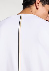 Biele tričko má okrúhly výstrih a zadný dizajn s vertikálnymi čiernymi a zlatými pruhmi zdôrazňujúcimi stredný šev. Hladká textúra.