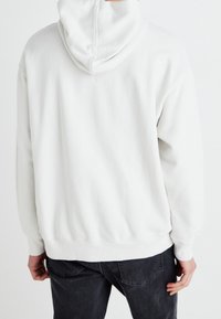 Světlý šedý mikinový svetr s kapucí, měkký materiál, pohodlný střih, žebrované manžety a lem, viditelné švy na kapuci, nošený s tmavě šedými kalhotami.