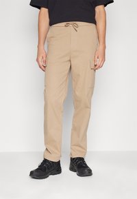 Man in beige cargobroeken met een trekkoord in de taille, zwarte sneakers en een zwart t-shirt met korte mouwen, staande tegen een witte achtergrond.