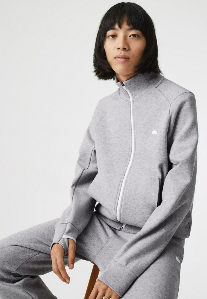 Chándal atlético gris hecho de tela suave, con un alto cuello, cierre de cremallera y un pequeño logo. Los pantalones tienen un diseño ajustado con puños en los extremos.