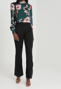 Blusa con stampa floreale verde e fiori rosa, colletto alto e maniche lunghe, abbinata a pantaloni neri a vita alta e sandali con cinturini.