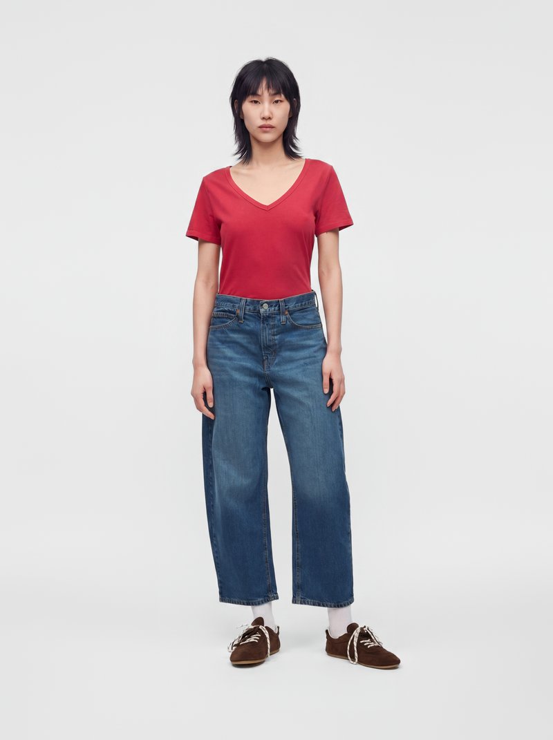 Femme debout face à l'avant portant un T-shirt rouge à col en V, un jean bleu à jambes larges, des chaussettes blanches et des chaussures brunes sur un fond uni.