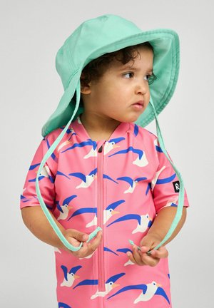 Małe dziecko noszące miętowy kapelusz przeciwsłoneczny i różową bluzę na zamek z niebieskimi i białymi nadrukami ptaków, patrzy na bok, trzymając sznurki od kapelusza.