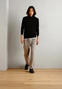 Czarny sweter z długim rękawem, jasnoszare spodnie i czarne buty. Model stoi przed gładką ścianą, podkreślając prostotę stylizacji.
