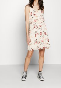 Šaty s květinovým potiskem z krémové tkaniny, mají véčkový výstřih, nastavitelné ramínka a elastický pas. Vytvořeno s černými vysokými teniskami.