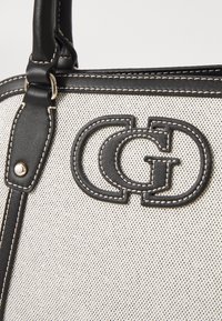 Sac fourre-tout noir et blanc avec une surface en toile texturée, présentant un logo noir en cuir bien en vue, du matériel en acier inoxydable et des coutures blanches contrastantes.