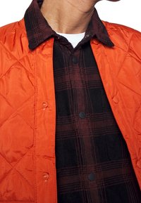 Quiltad orange jacka med snap-stängning och svarta knappar, lager på lager över en mörk rutig skjorta. Har kontrasterande texturer och färger.