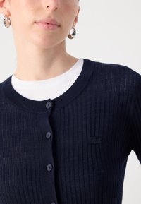 Marinblå ribbad cardigan med rund hals, knappar framtill och en liten broderad logotyp på vänster sida. Mjukt, tunt stickat tyg.