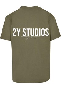 Olīvu zaļš kokvilnas T-krekls ar izteiksmīgu balto tekstu "2Y STUDIOS" un mazāku tekstu "TIKAI PAGAIDU LOCEKĻIEM" uz muguras. Vienkāršs dizains.