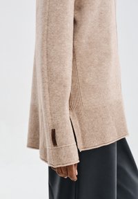 Beżowy sweter o miękkim, teksturowanym splocie z żeberkowymi akcentami na rękawach i dolnym wykończeniu. Posiada rozcięcia po bokach oraz małą metkę z logo.