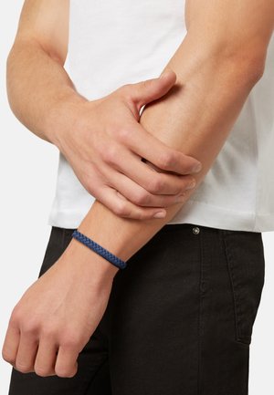Navy blue braided bracelet made of leather worn on a wrist, with a close-up view of hand resting on an arm. Simple design, textured finish.