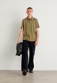 Polo en vert olive avec manches courtes, pantalon noir à jambes larges et veste en cuir noire drapée sur le bras gauche. Texture de tissu lisse, coupe décontractée.