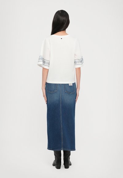 Bílý tričko s modrými vyšívanými detaily na rukávech, spojené s tmavými džínsovými maxi sukněmi. Model má na sobě černé kotníkové boty.