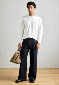 Balta krekla ar garām piedurknēm, tumšas taisnas džinsa bikses un melni apavi. Vienā rokā tiek turēta brūna soma ar īsu rokturi.