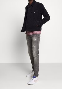 Chaqueta de pana negra con dos bolsillos en el pecho, superpuesta sobre una camisa de rayas burdeos y blanca. Jeans desgastados en gris claro y zapatillas blancas.