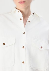 Hvit skjorte med knappelukking laget av denim, med to brystlommer, messingknapper og klassisk kragedesign. Teksturert stoffdetalj synlig.