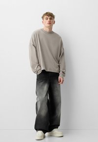 Světle šedá oversized košile s dlouhým rukávem v kombinaci s černými vybledlými širokými džínami a bílými teniskami. Hladká tkanina a uvolněný střih.