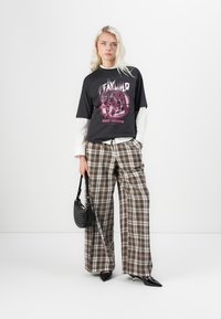 Schwarzes Grafik-T-Shirt mit Leopardenmuster, über ein weißes Langarmshirt geschichtet. Braune karierte weitgeschnittene Hose und eine schwarze strukturierte Handtasche.