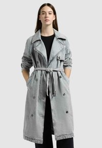 Frau trägt einen hellgrauen, mit Gürtel versehenen Trenchcoat über einem schwarzen Outfit und steht mit den Händen in den Taschen vor einem schlichten Hintergrund.